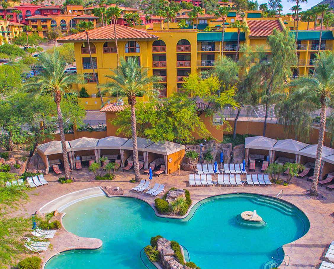Aerial view of the Grotto Pool at The Falls Water Village with surrounding cabanas, lounge chairs and palm trees and resort buildings and mountains in the background