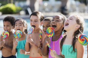 A group of six multi-ethnic children standing together at a swimming pool on a sunny day, licking giant lollipops. The children are 6 to 9 years old. Multi-ethnic children having fun licking lollipops by swimming pool