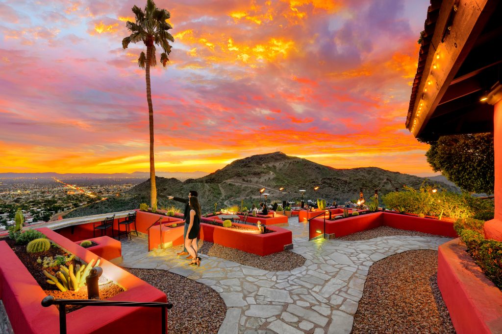 Couple on outdoor patio with views of surrounding mountains, a fiery sunset sky and city lights