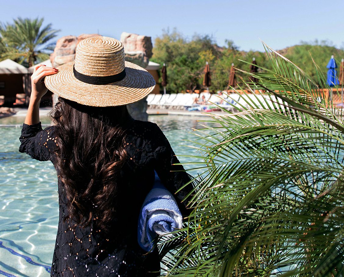 Woman strolling next to the pool with palm trees, cabanas and mountains in the background