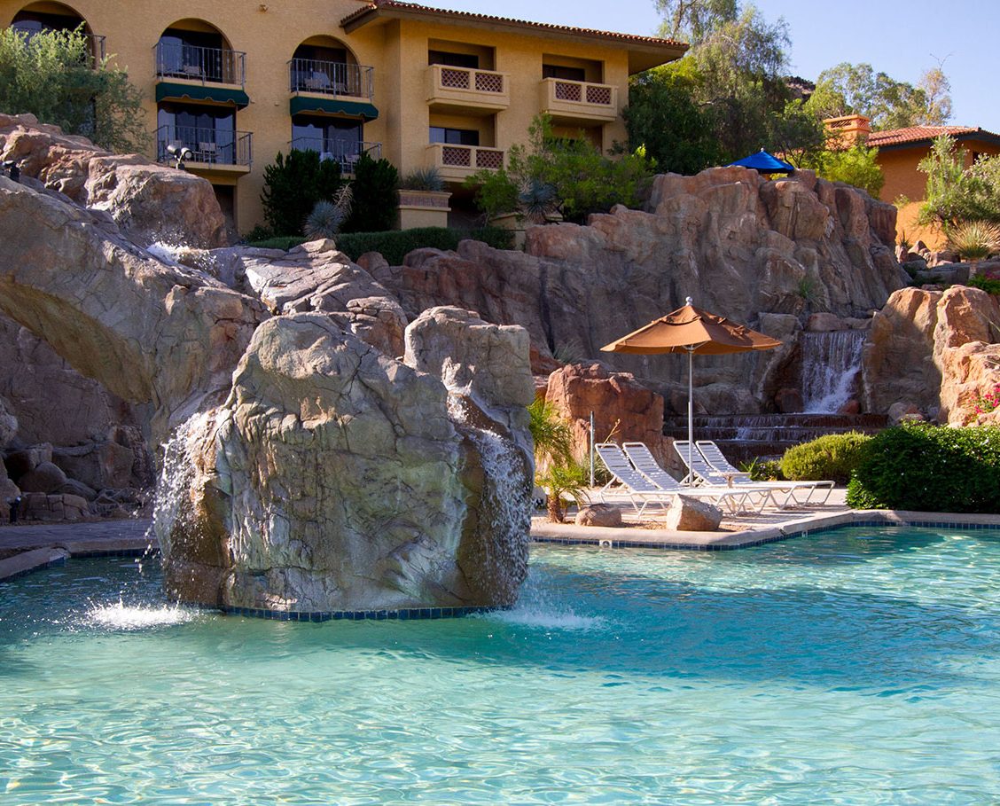 View sparkling pool, waterfalls, sun-soaked lounge chairs and a resort building in the background