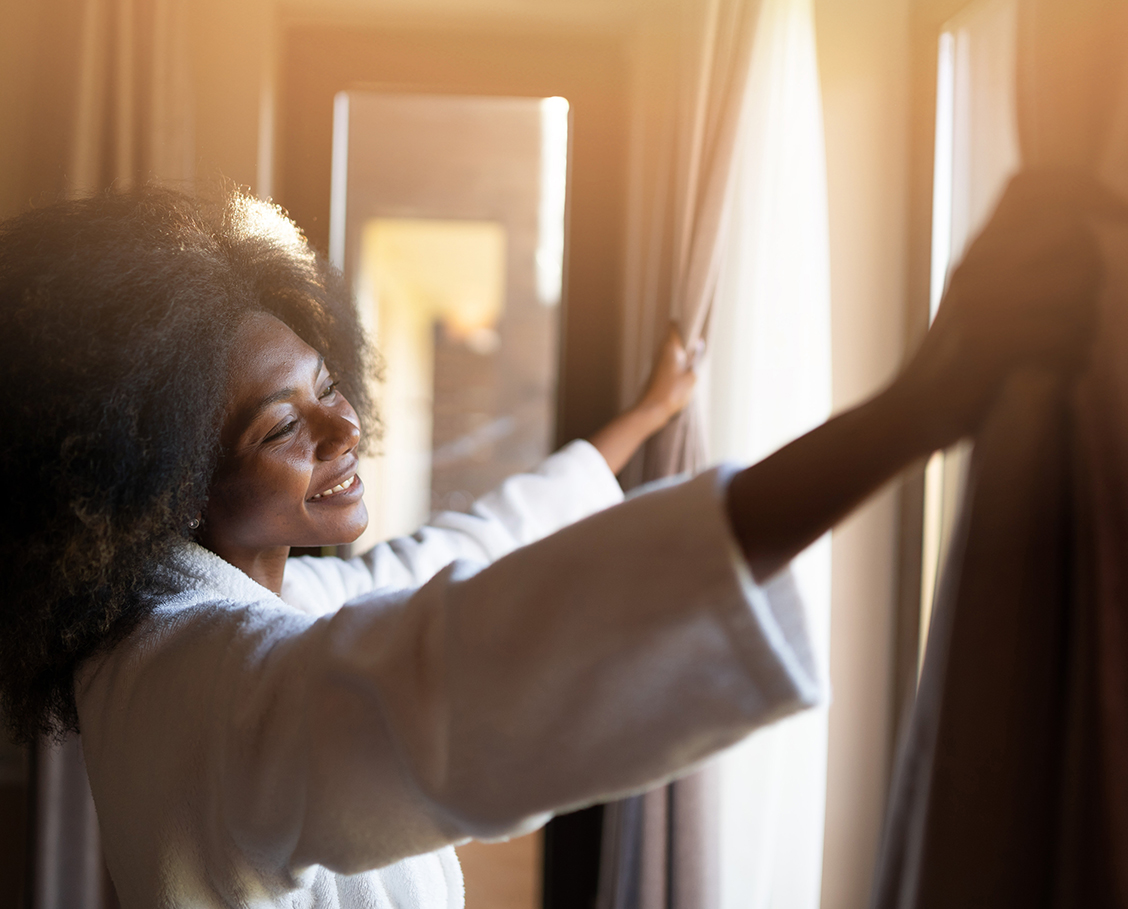 African woman pulling the curtains in hotel room, morning.